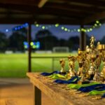 Trophies and medals with blue and lime ribbons on a table at the Bonnet Bay FC clubhouse at dusk, floodlit pitch behind