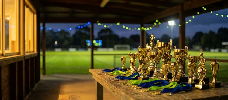 Trophies and medals with blue and lime ribbons on a table at the Bonnet Bay FC clubhouse at dusk, floodlit pitch behind